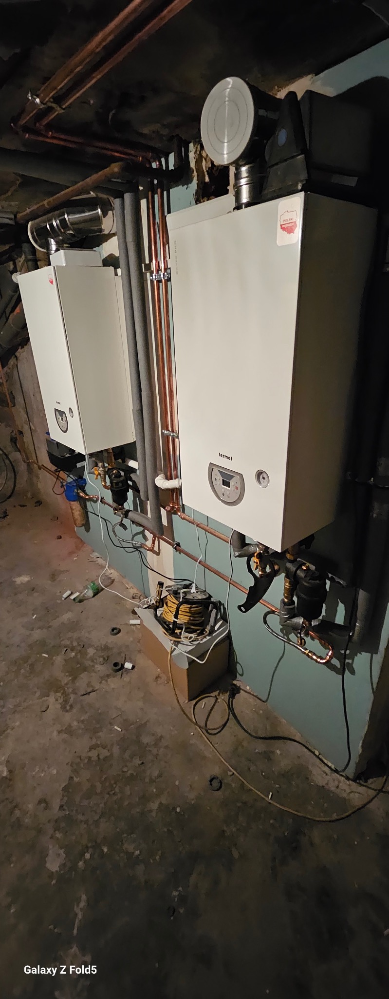 Dwa białe piece gazowe Termet zamontowane na ścianie z miedzianymi rurami. Widoczne przewody i akcesoria instalacyjne. Podłoga betonowa.