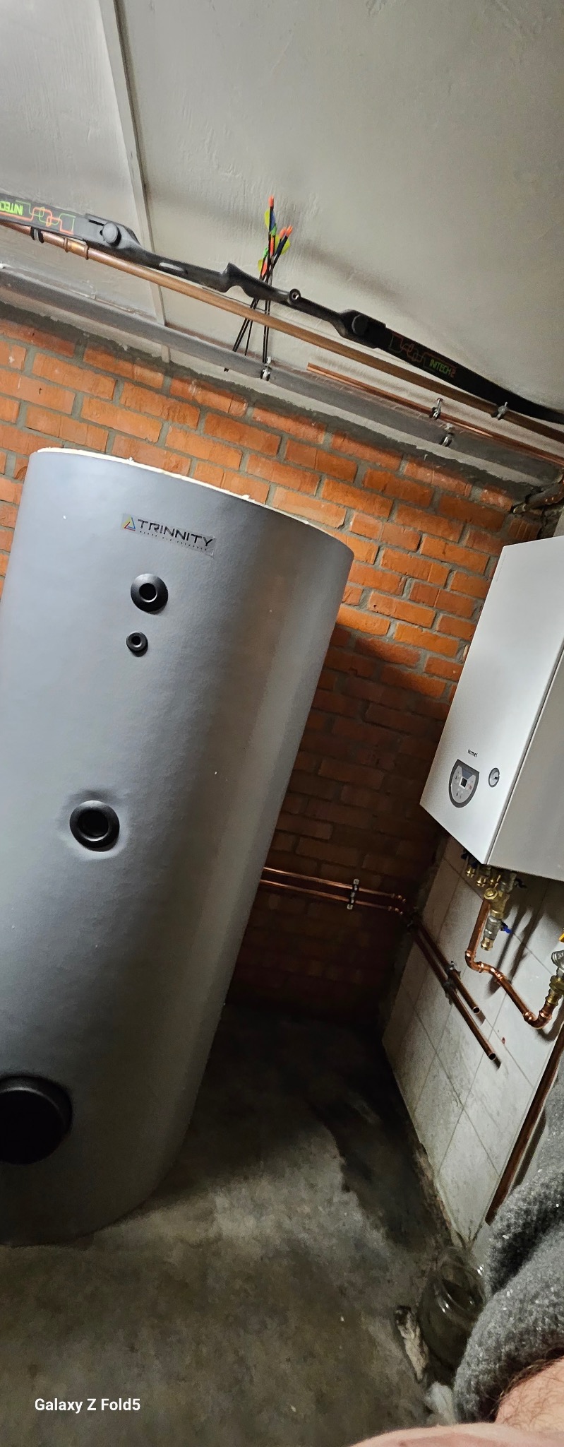 Szary zasobnik ciepłej wody użytkowej Trinity obok białego pieca gazowego Fermet, z miedzianymi rurami i ceglaną ścianą w tle. Widoczny łuk sportowy na ścianie.