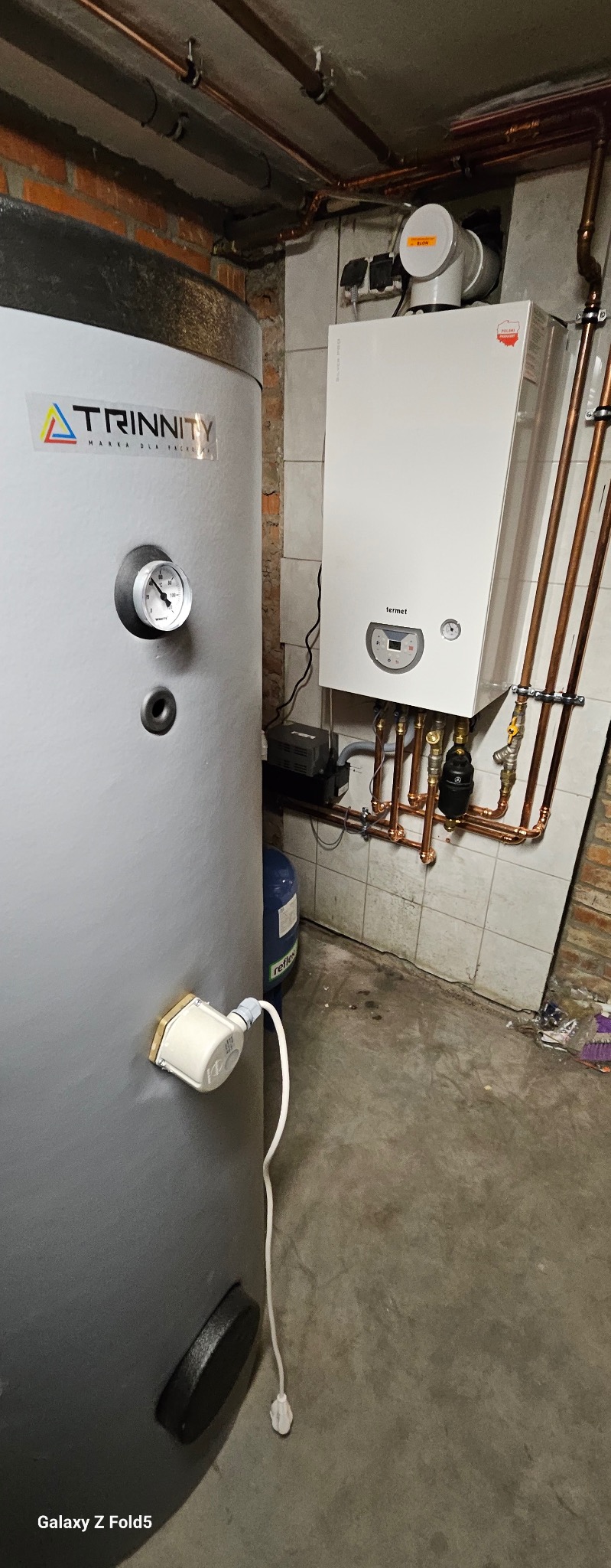 Szary zasobnik ciepłej wody Trinity i biały piec gazowy Termet z miedzianą instalacją w kotłowni z częściowo wykończonymi ścianami z cegły i płytek.