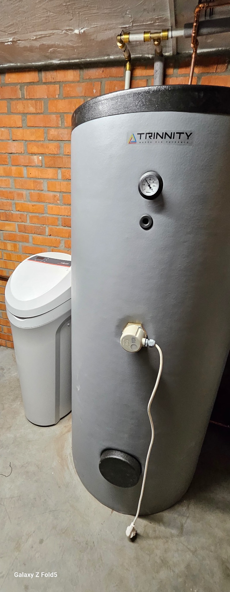 Szary zbiornik ciepłej wody użytkowej marki Trinnity z manometrem i podłączeniem elektrycznym, obok zmiękczacz wody, na tle ceglanej ściany.