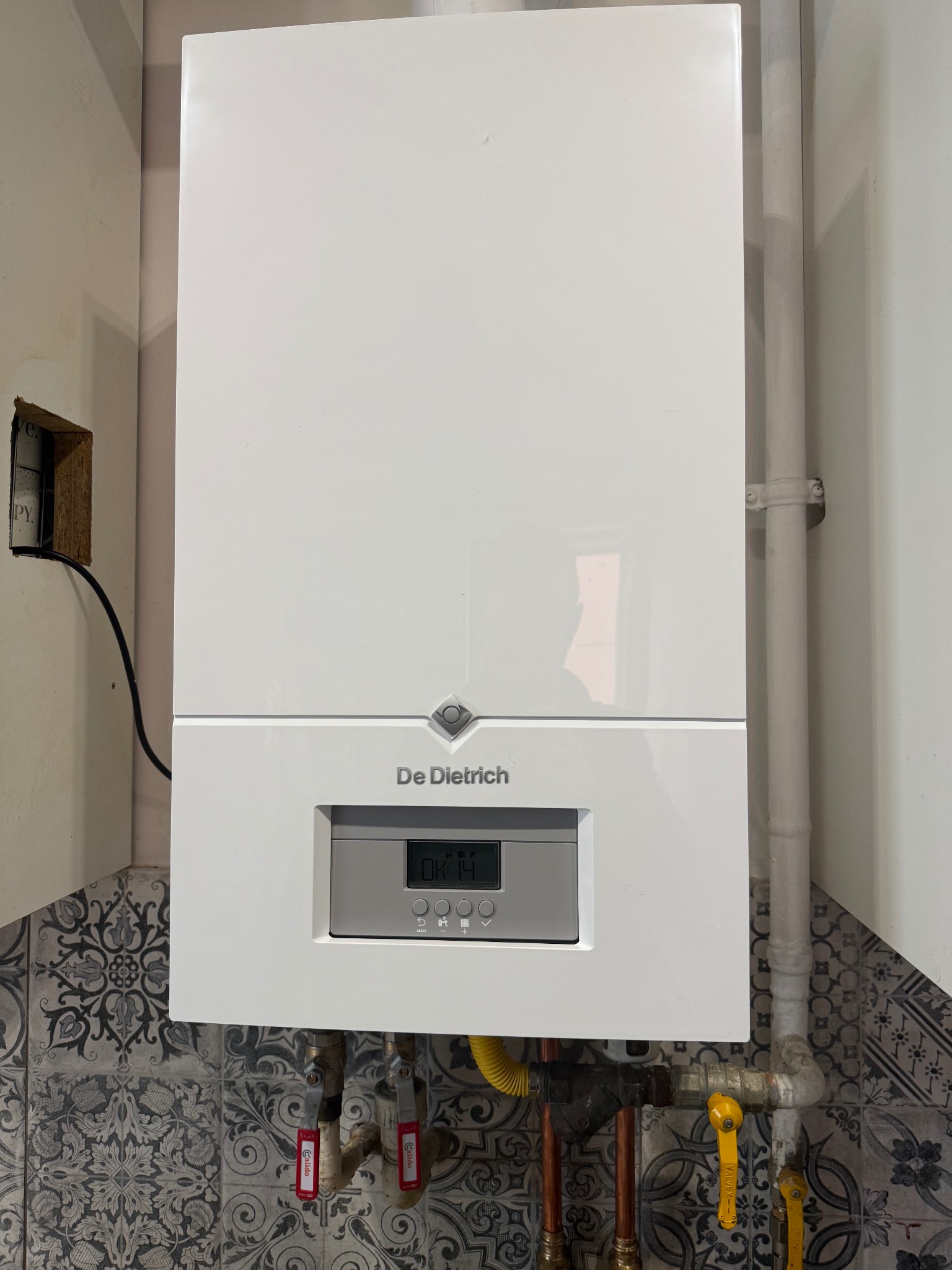 Biały, wiszący piec gazowy De Dietrich na tle wzorzystych płytek. Widoczne rury i zawory. Minimalistyczny design urządzenia wkomponowany w domową przestrzeń.