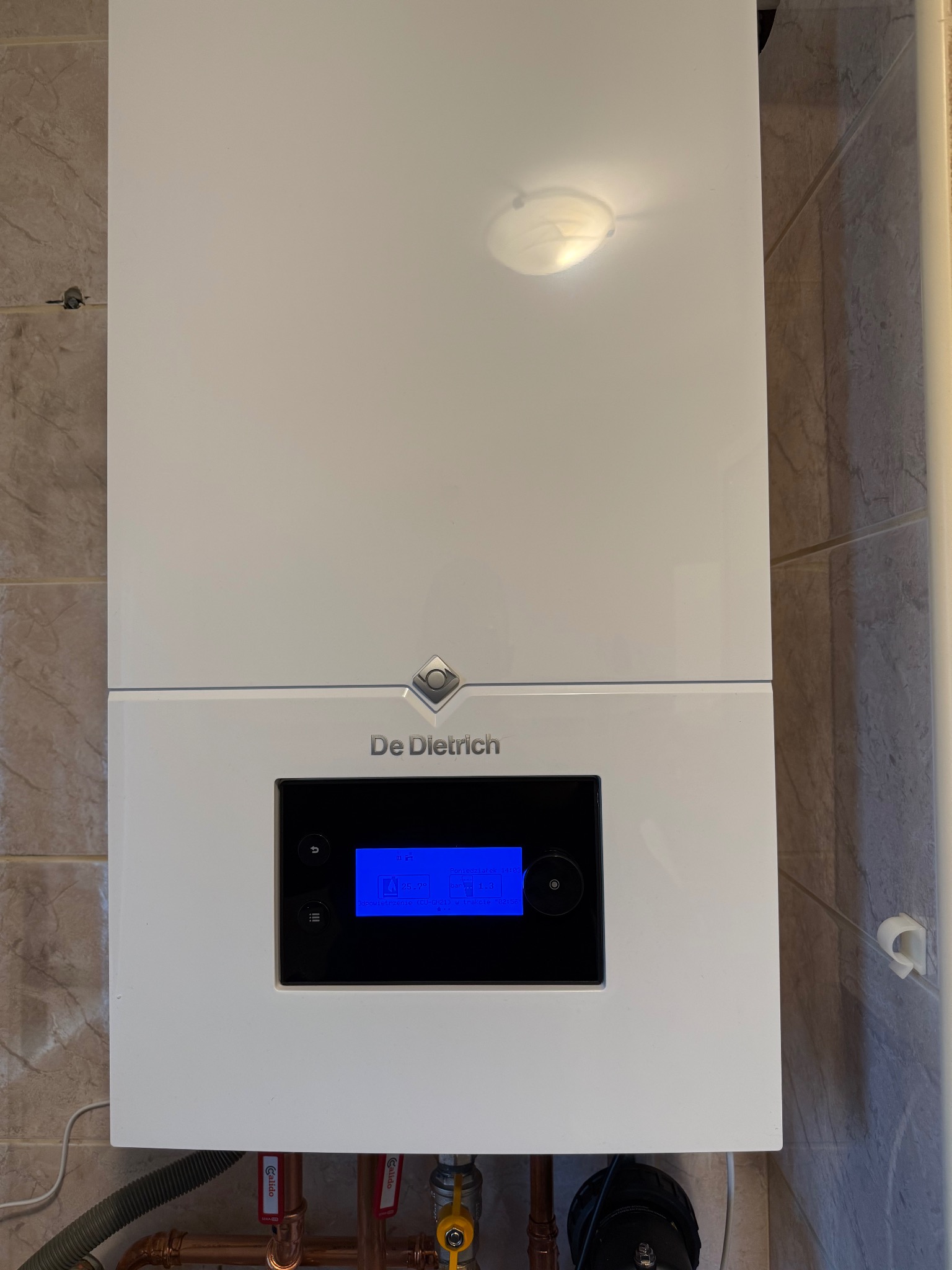 Biały, wiszący piec gazowy De Dietrich na tle beżowych płytek. Widoczne miedziane rury i zawory. Na wyświetlaczu pieca widoczny niebieski ekran z informacjami.