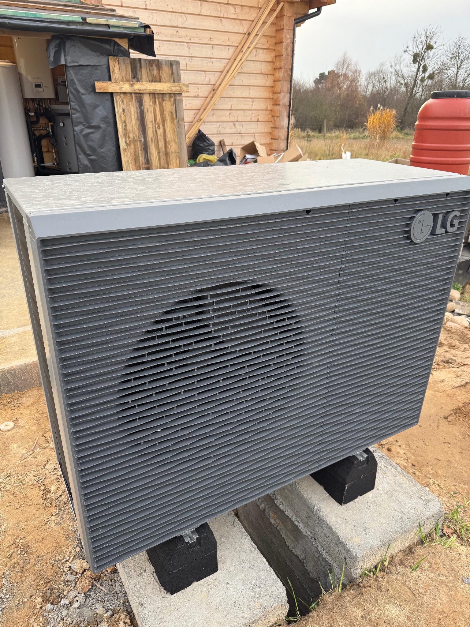 Szara pompa ciepła LG, zamontowana na betonowych blokach, z widocznym budynkiem w tle. Instalacja zewnętrzna w trakcie prac wykończeniowych.