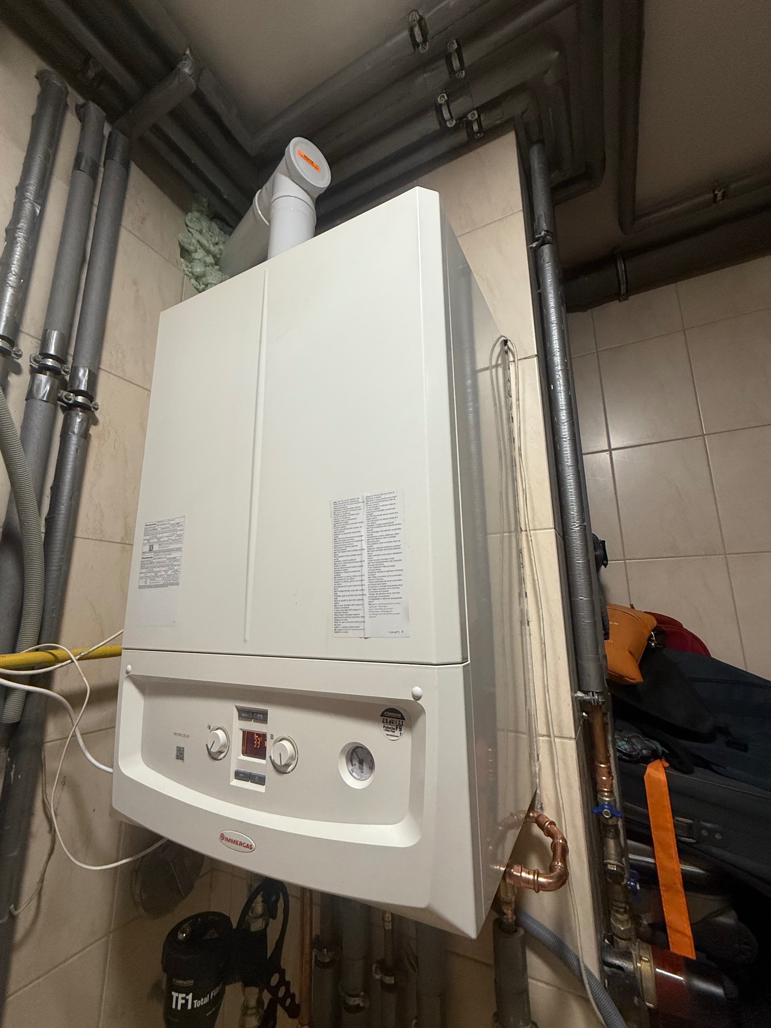 Biały piec gazowy Immergas z widocznymi rurami instalacji, zamontowany na kafelkowanej ścianie w pomieszczeniu technicznym. Widoczne elementy instalacji i uszczelnienia.