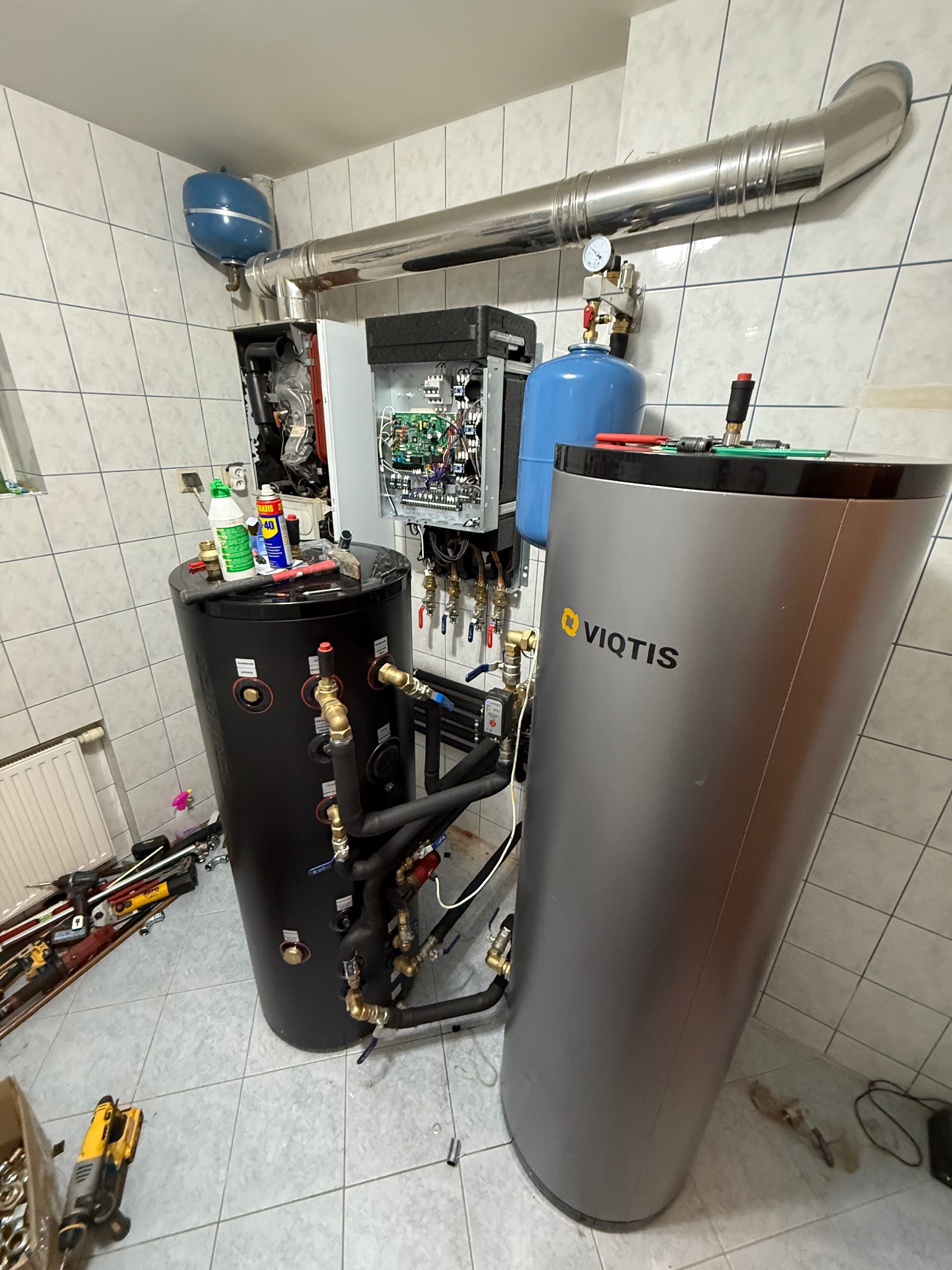 Instalacja kotłowni z dwoma zbiornikami, czarnym i srebrnym Viqtis, oraz widoczną instalacją hydrauliczną i narzędziami na podłodze w pomieszczeniu wyłożonym kafelkami.