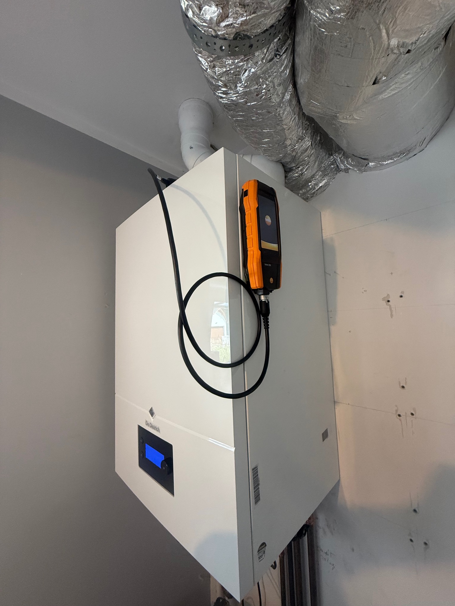 Biały piec gazowy De Dietrich z podłączonym analizatorem spalin Testo 300, widoczne rury wentylacyjne. Ściana z śladami po montażu.