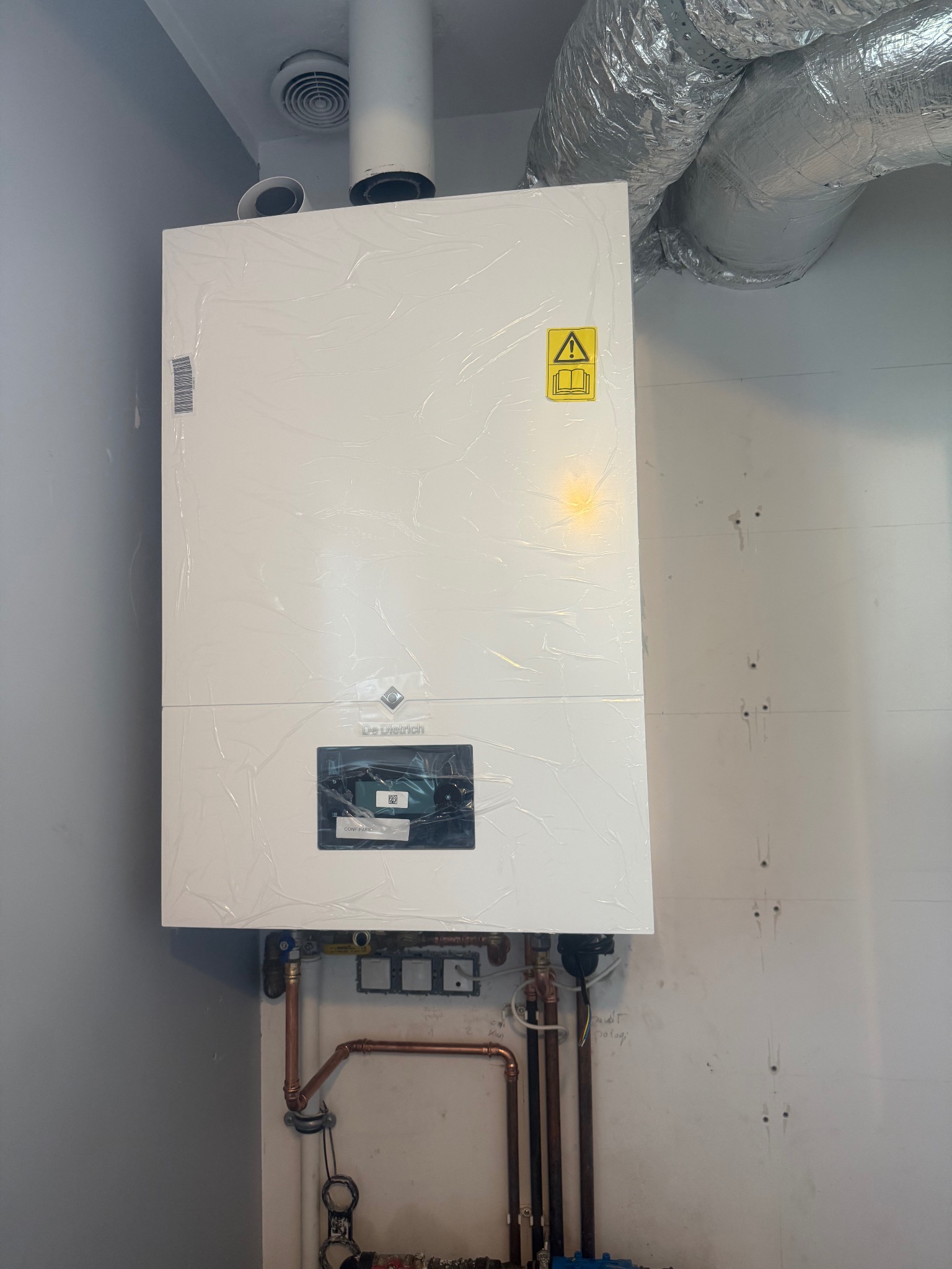 Nowy, biały piec gazowy De Dietrich, jeszcze w folii ochronnej, zamontowany na ścianie z widocznymi rurami miedzianymi i instalacją. Górą srebrny przewód wentylacyjny.