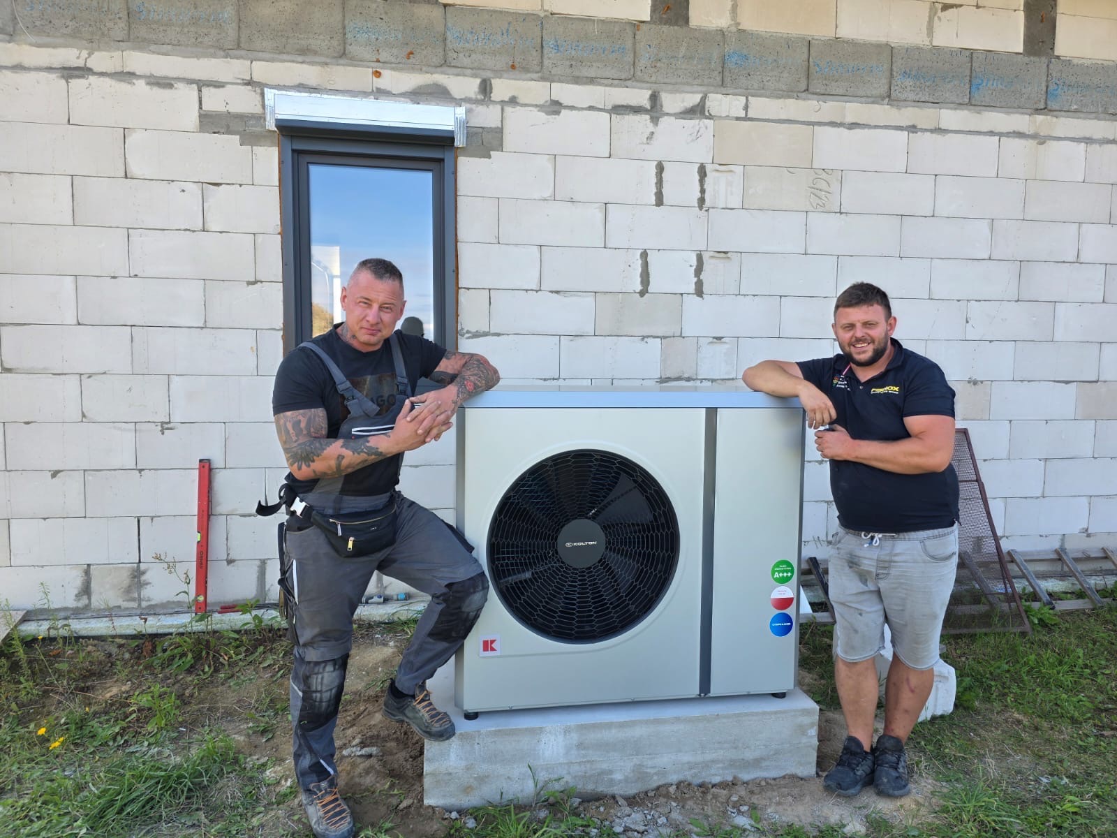 Dwóch instalatorów pozuje obok zamontowanej pompy ciepła Kolton na betonowym fundamencie, na tle ściany budynku z białego bloczka i okna z roletą.