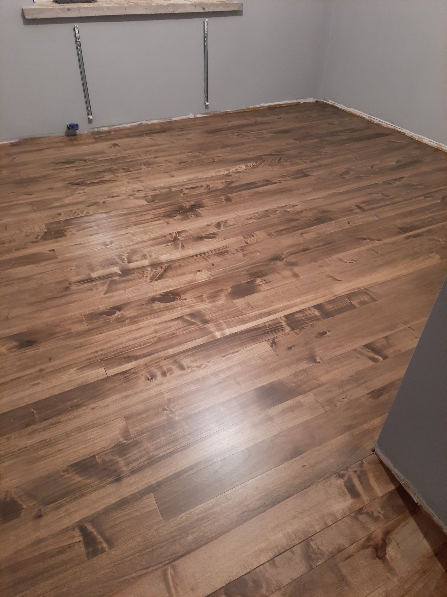 Ułożony parkiet z wyraźnym usłojeniem w odcieniach brązu, widoczne listwy przypodłogowe oraz metalowe wsporniki na ścianie.