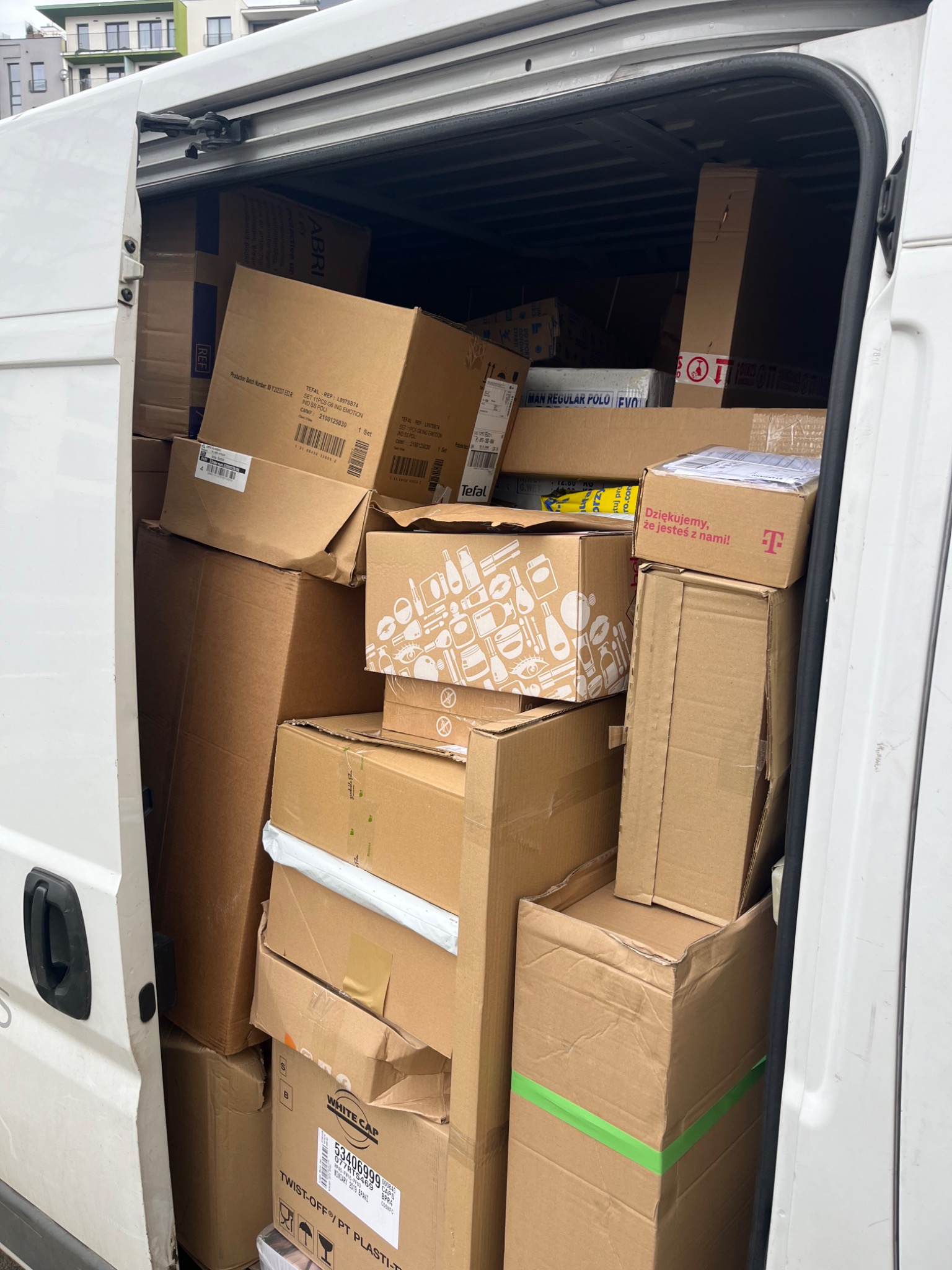 Wnętrze białego busa wypełnione po brzegi kartonami różnych rozmiarów, niektóre z logo firm takich jak Tefal i T-Mobile, zabezpieczone taśmami.