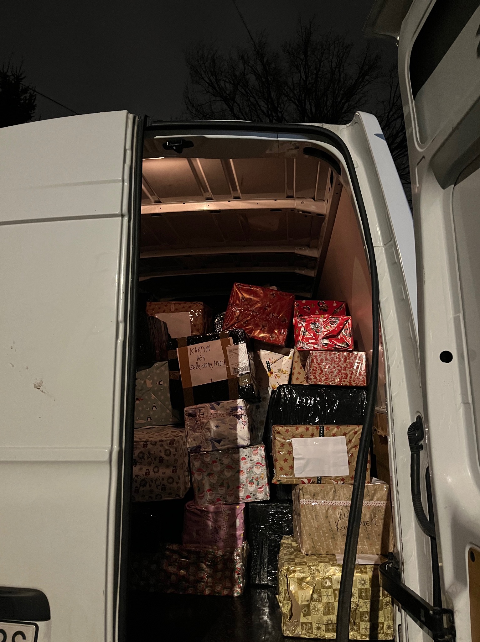 Wnętrze białego busa załadowane po brzegi zapakowanymi prezentami świątecznymi, widoczne etykiety adresowe, ujęcie nocne.