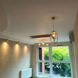 Wykończone wnętrze pokoju: kremowe ściany, biały sufit z oświetleniem punktowym i wiszącą lampą. Widok na okno z zielenią na zewnątrz i grzejnik pod nim.