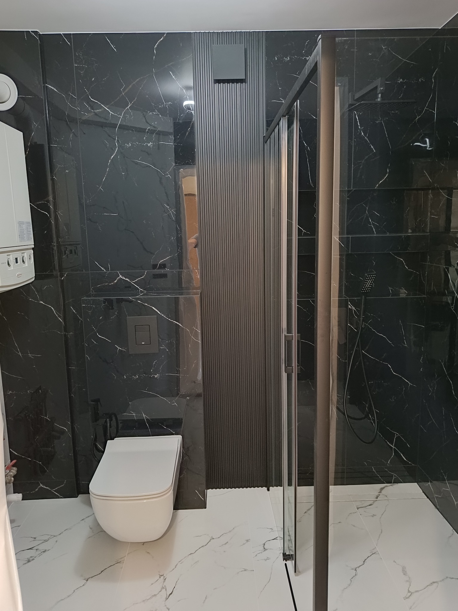 Nowoczesna łazienka z czarnymi marmurkowymi płytkami na ścianach, białą podłogą z marmurowym wzorem i kabiną prysznicową z ciemną ramą. Wisząca toaleta i grzejnik na ścianie.
