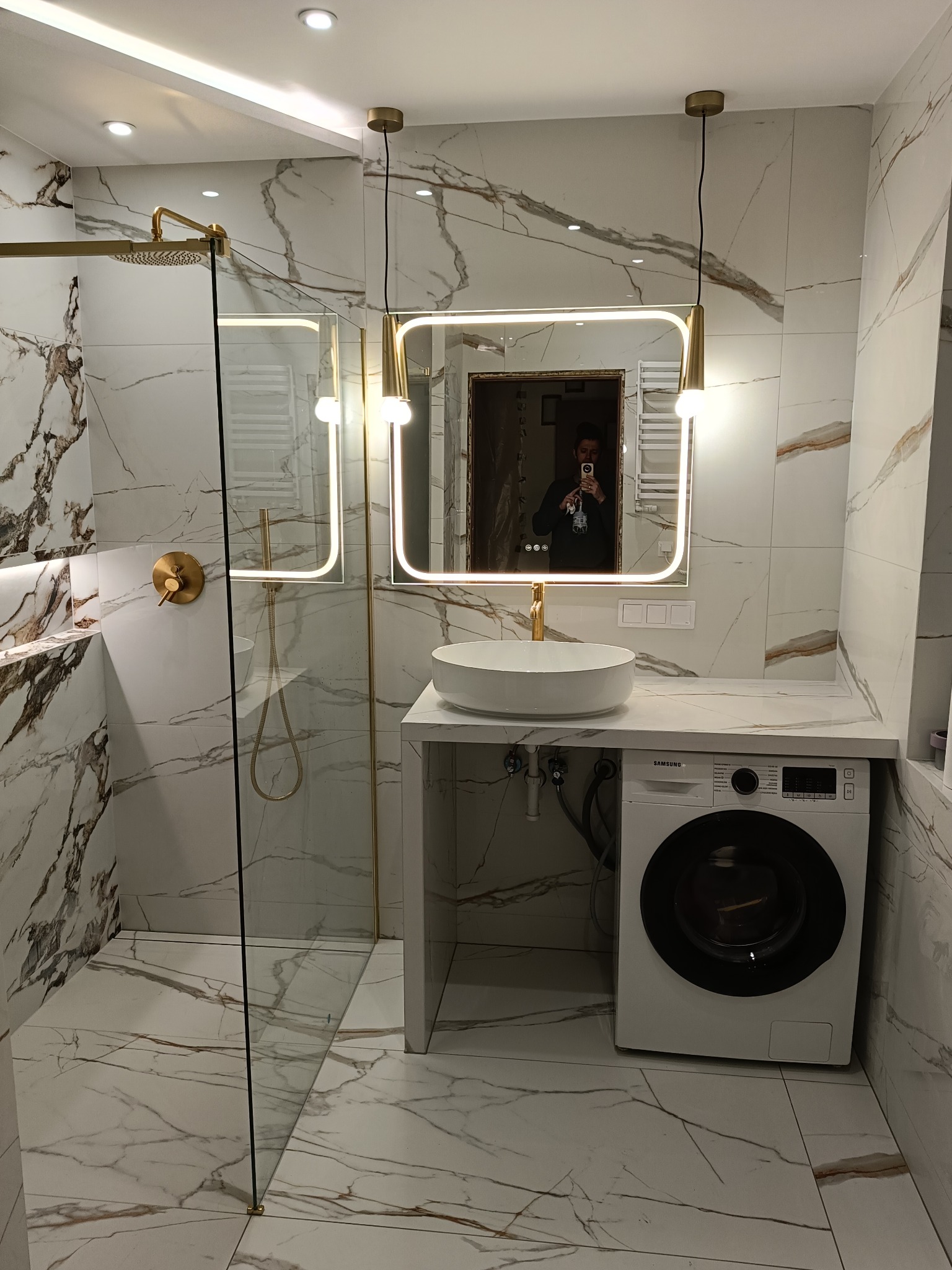 Wykończona łazienka w bieli i złocie: kabina prysznicowa typu walk-in, umywalka nablatowa, pralka pod blatem. Marmurowe płytki, oświetlenie LED.