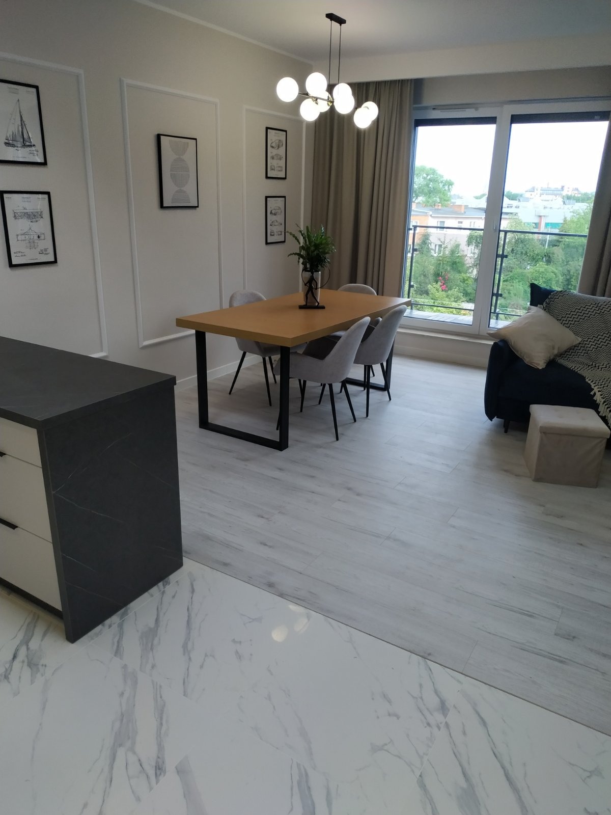 Jasne wnętrze: stół z drewnianym blatem i szarymi krzesłami, minimalistyczne dekoracje ścienne, okno z widokiem na zieleń, marmurowe płytki na podłodze.