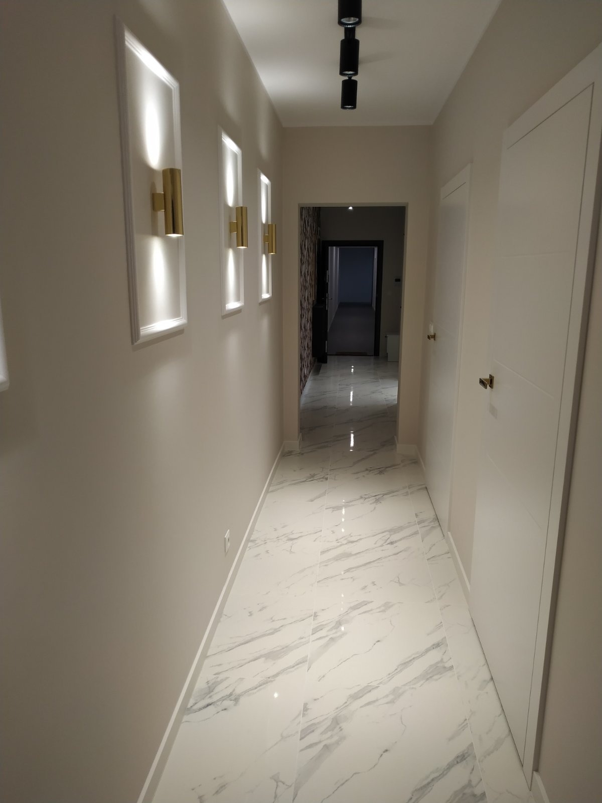 Wykończony korytarz z białymi drzwiami, marmurową podłogą i dekoracyjnym oświetleniem ściennym w złotych oprawach. Widok w głąb mieszkania.