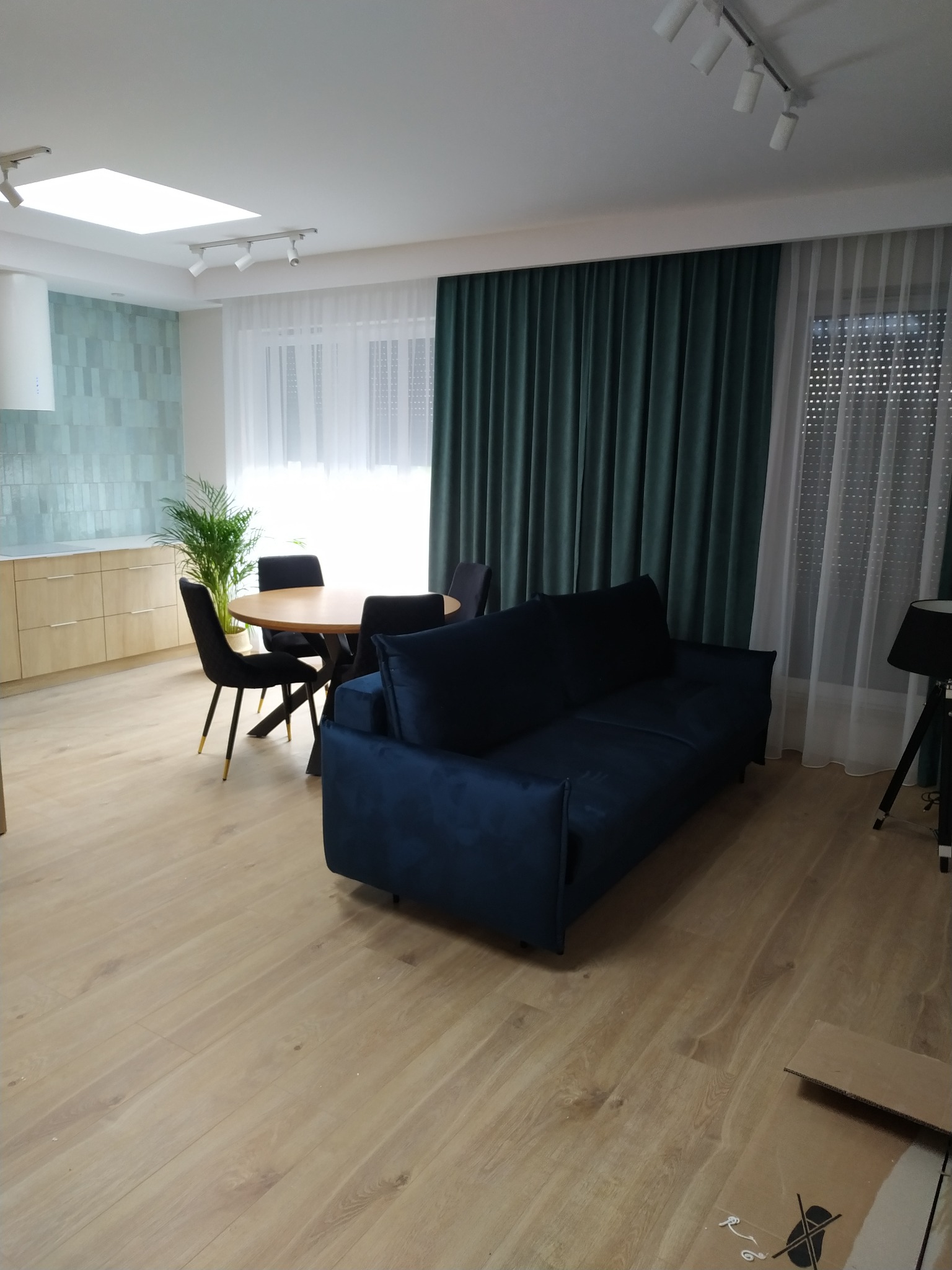 Nowoczesne wnętrze: błękitna sofa, okrągły stół z krzesłami, drewniana podłoga, zielone zasłony i akcenty turkusowych płytek w kuchni.