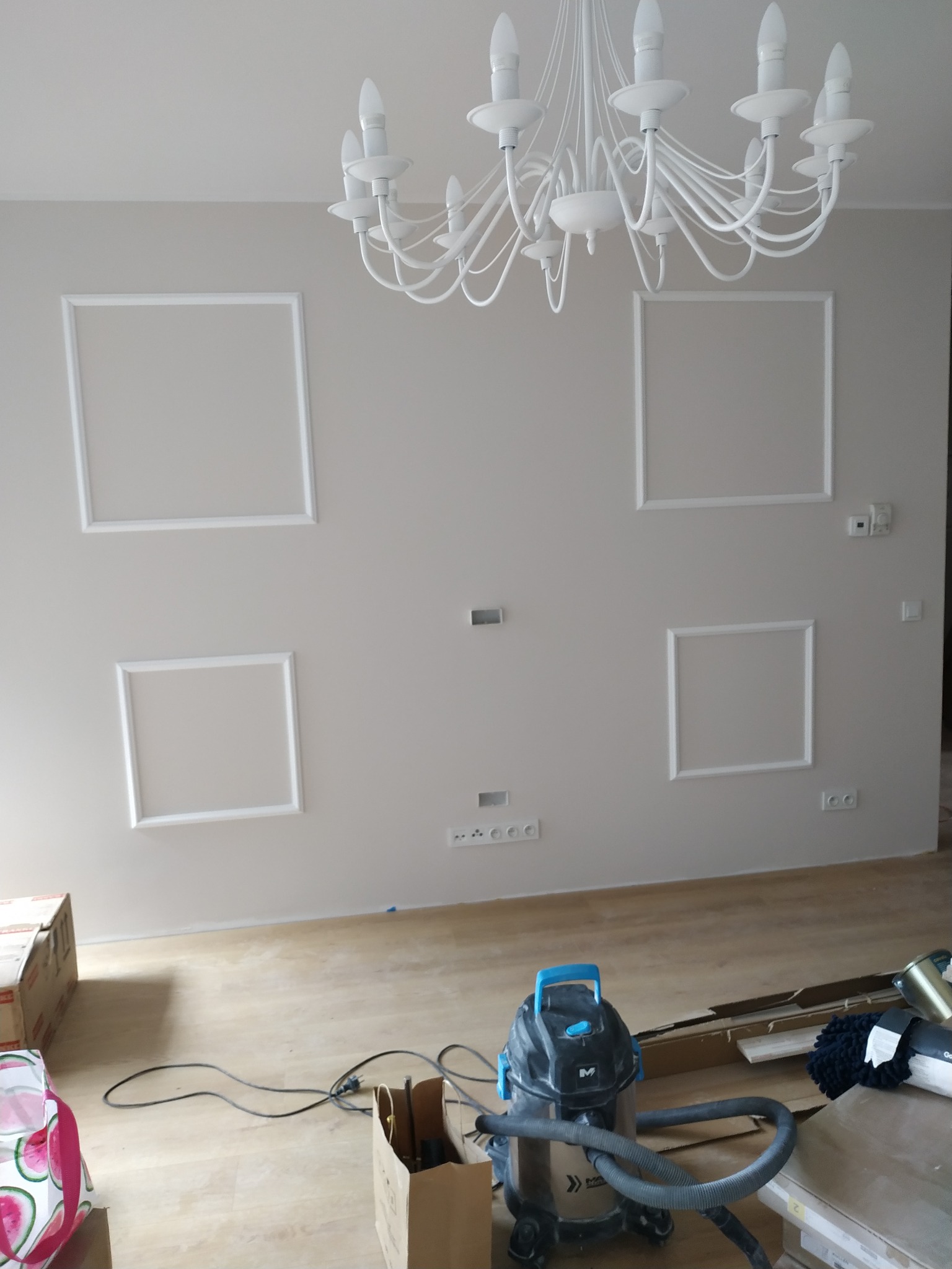 Ściana w odcieniach beżu z białymi, kwadratowymi ramkami i ozdobnym żyrandolem. Na podłodze widoczny odkurzacz przemysłowy i kartony.
