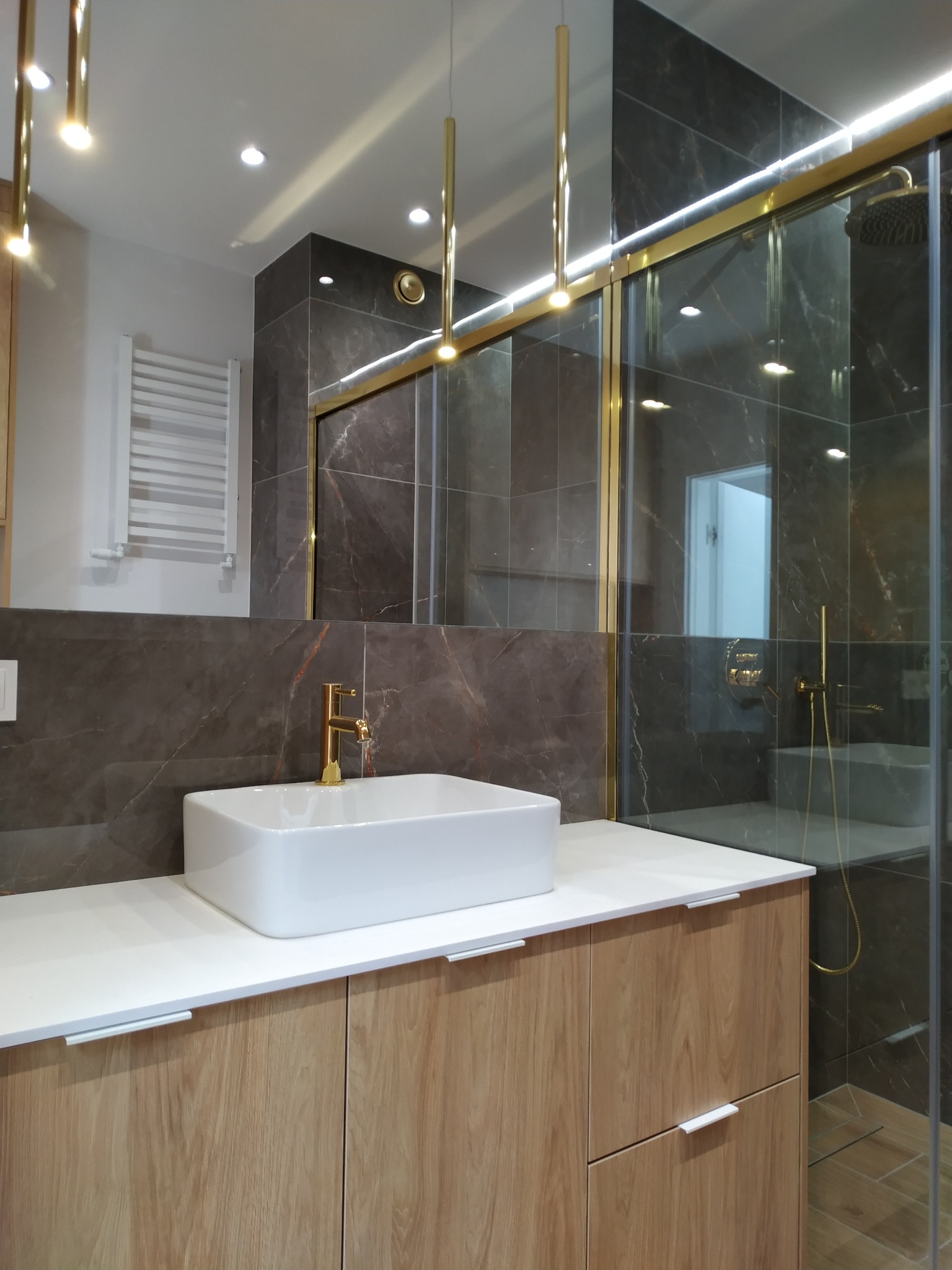 Elegancka łazienka z umywalką nablatową, złotą armaturą i kabiną prysznicową w ciemnych płytkach imitujących marmur. Nowoczesne oświetlenie.
