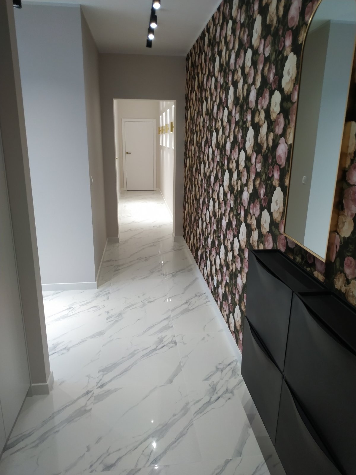 Wykończony korytarz z białymi marmurowymi płytkami, tapetą w kwiaty i czarną szafką na buty. Nowoczesne oświetlenie sufitowe i lustro w złotej ramie.