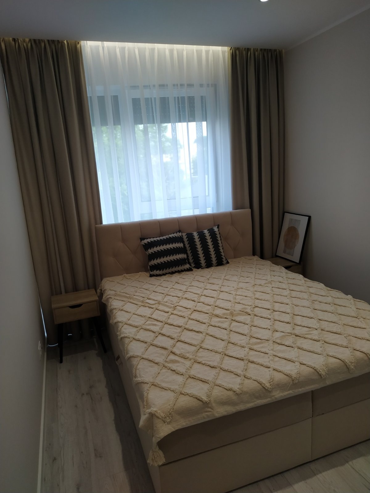 Beżowa sypialnia z łóżkiem, wzorzystymi poduszkami i zasłonami. Na ścianie minimalistyczny obraz. Jasne, subtelne oświetlenie.