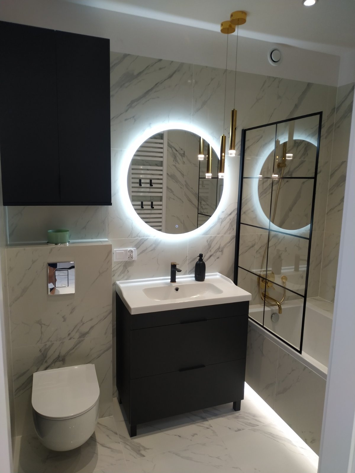 Nowoczesna łazienka z umywalką na szafce w kolorze grafitu, okrągłym lustrem z podświetleniem LED i kabiną prysznicową z czarnymi szprosami.