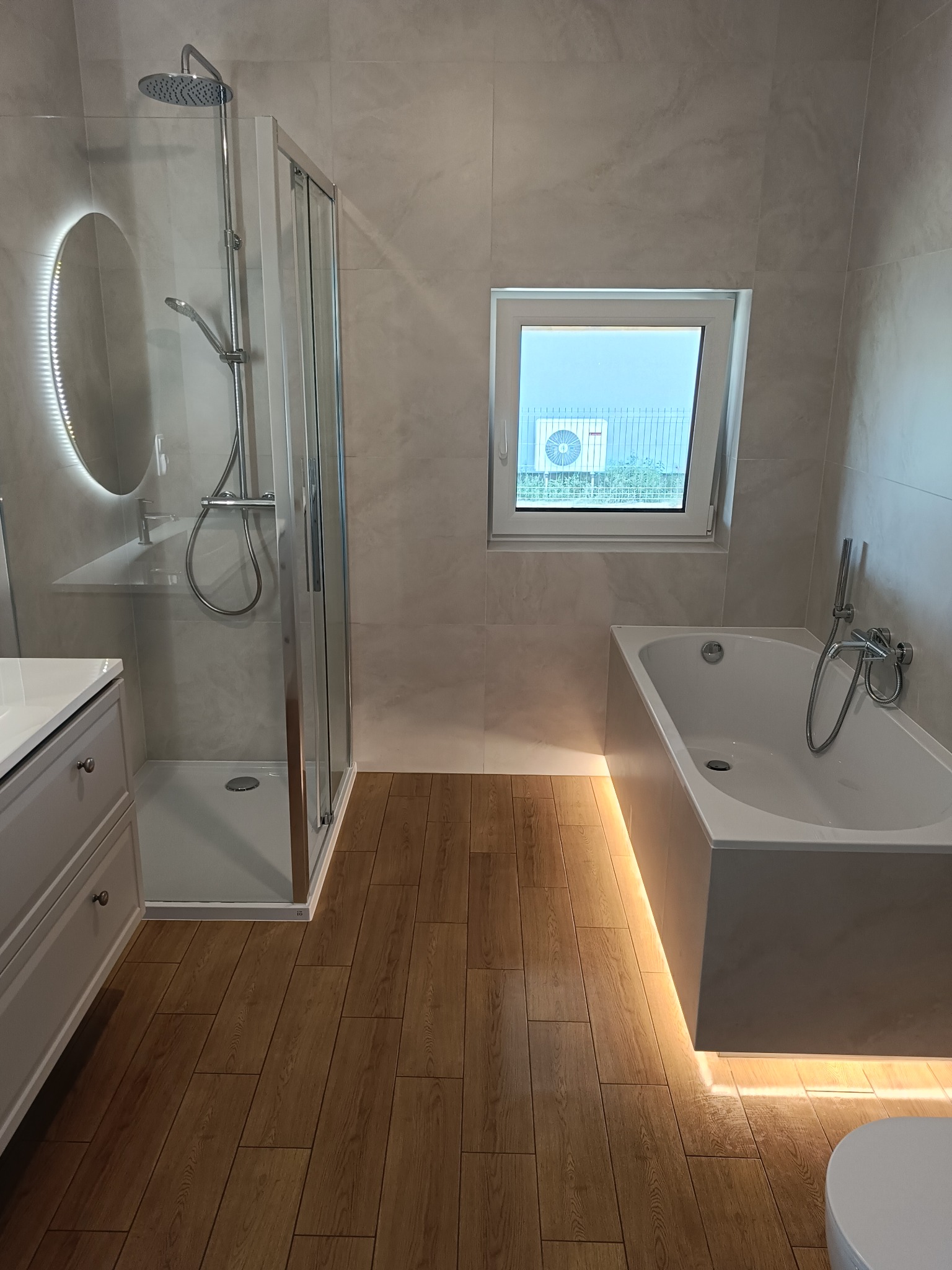 Nowoczesna łazienka z kabiną prysznicową typu walk-in, podświetlaną wanną i drewnopodobną podłogą. Minimalistyczny design, jasne płytki, okrągłe lustro z LED.