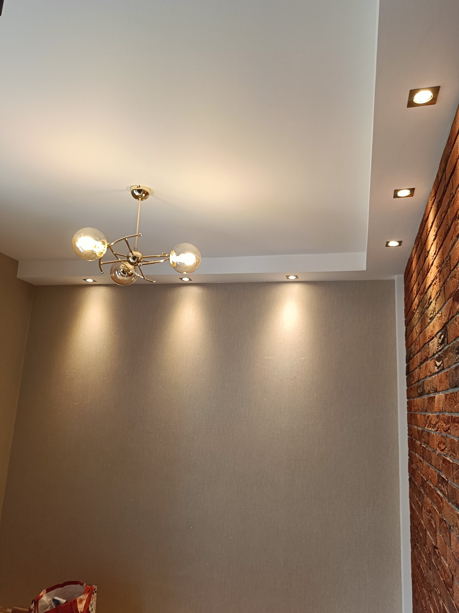 Beżowa ściana z połyskującą tapetą, nowoczesny żyrandol, sufit z halogenami i fragment ściany z cegły w wykończonym wnętrzu.