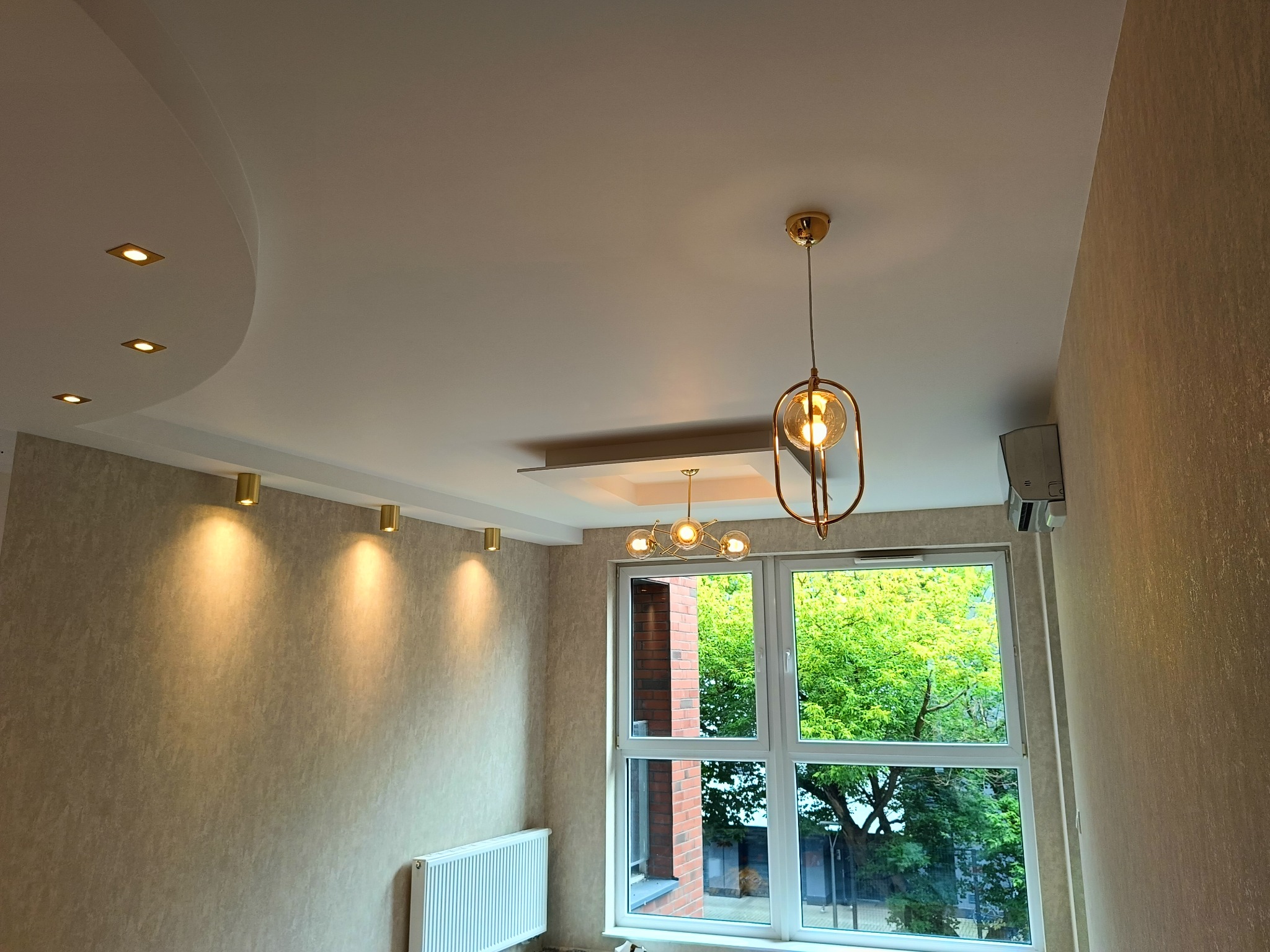 Wykończone wnętrze pokoju: kremowe ściany, biały sufit z oświetleniem punktowym i wiszącą lampą. Widok na okno z zielenią na zewnątrz i grzejnik pod nim.