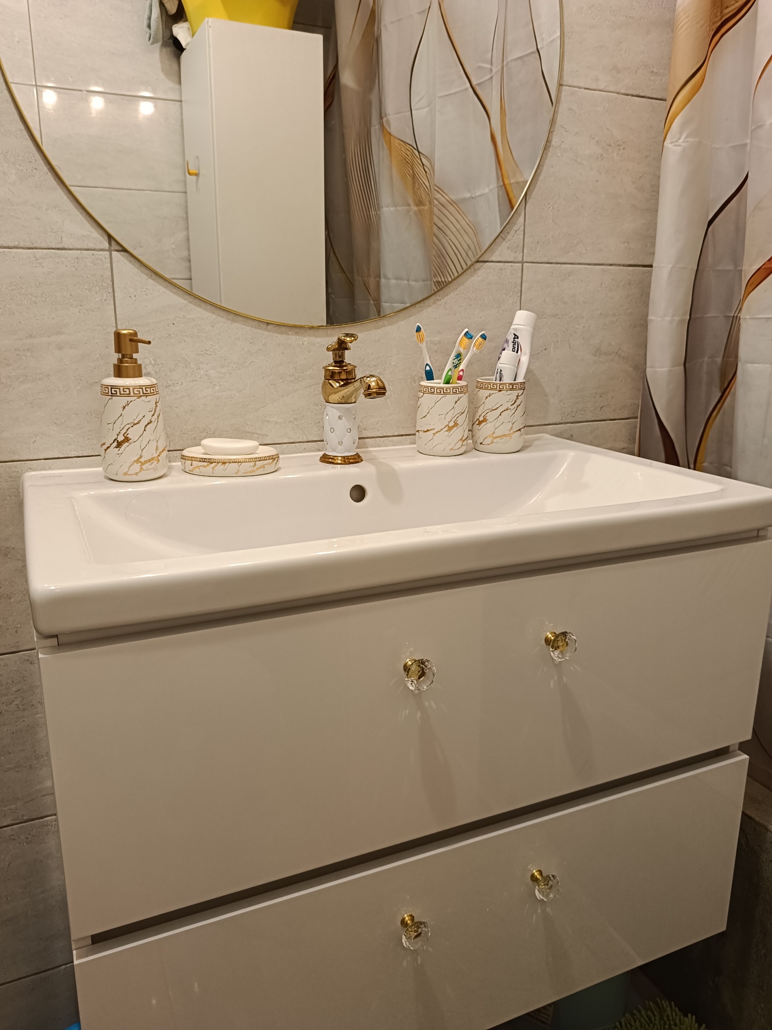 Biała szafka pod umywalkę z złotą armaturą i okrągłym lustrem w łazience z szarymi płytkami. Elegancki design z marmurkowymi akcentami.