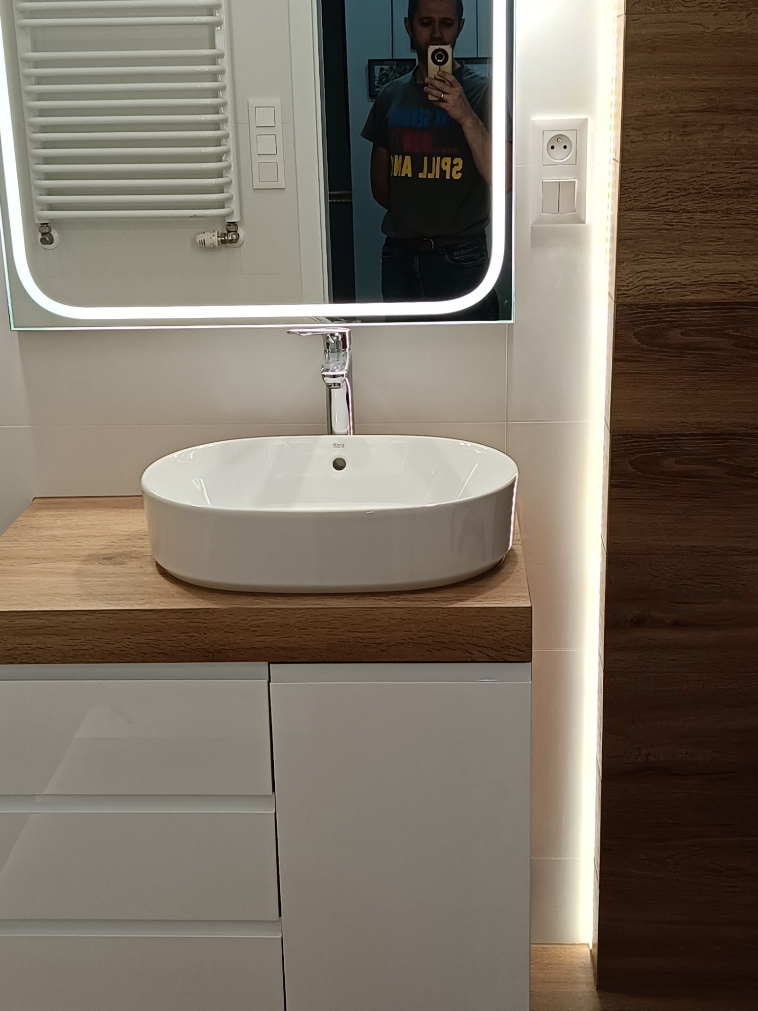 Nowoczesna łazienka: owalna umywalka Roca na drewnianym blacie, białe szafki, podświetlane lustro LED z odbiciem osoby, grzejnik drabinkowy i drewnopodobna okładzina.