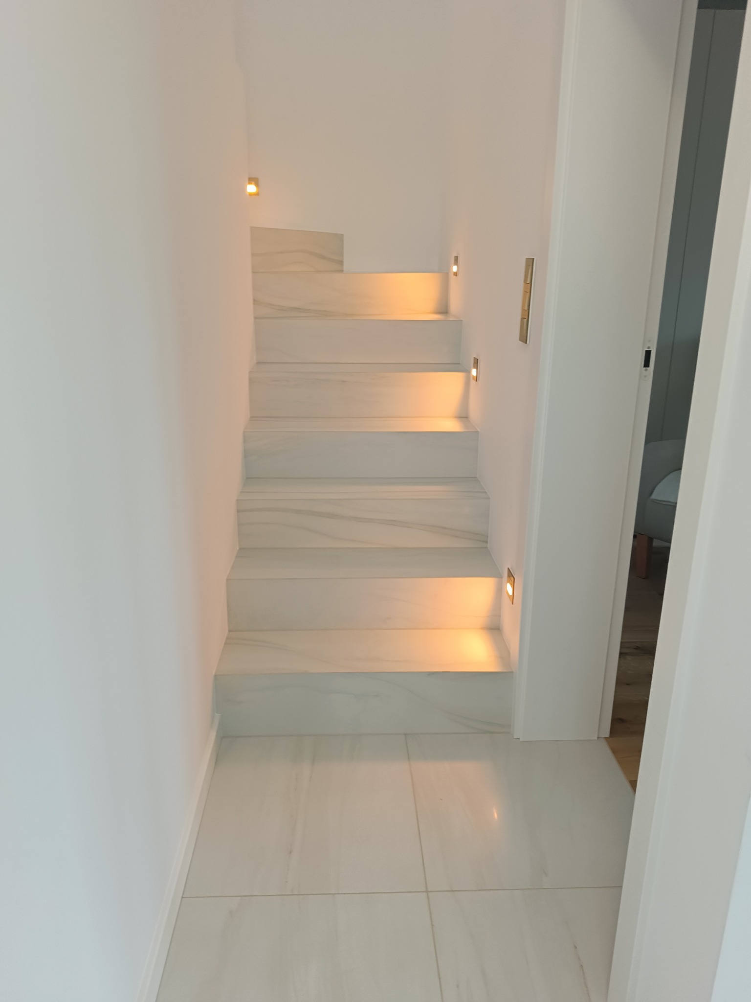 Białe marmurowe schody z subtelnym oświetleniem punktowym wzdłuż ściany, prowadzące w górę. Minimalistyczny design, widok z dołu.