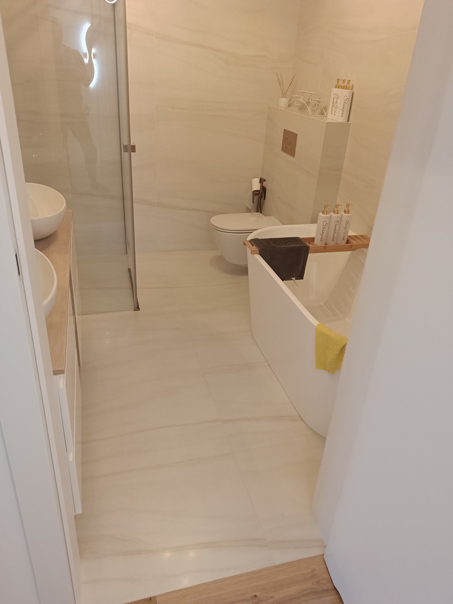 Jasna łazienka z wanną, umywalką i kabiną prysznicową. Płytki imitujące marmur na ścianach i podłodze. Minimalistyczny design z akcentami w kolorze drewna.