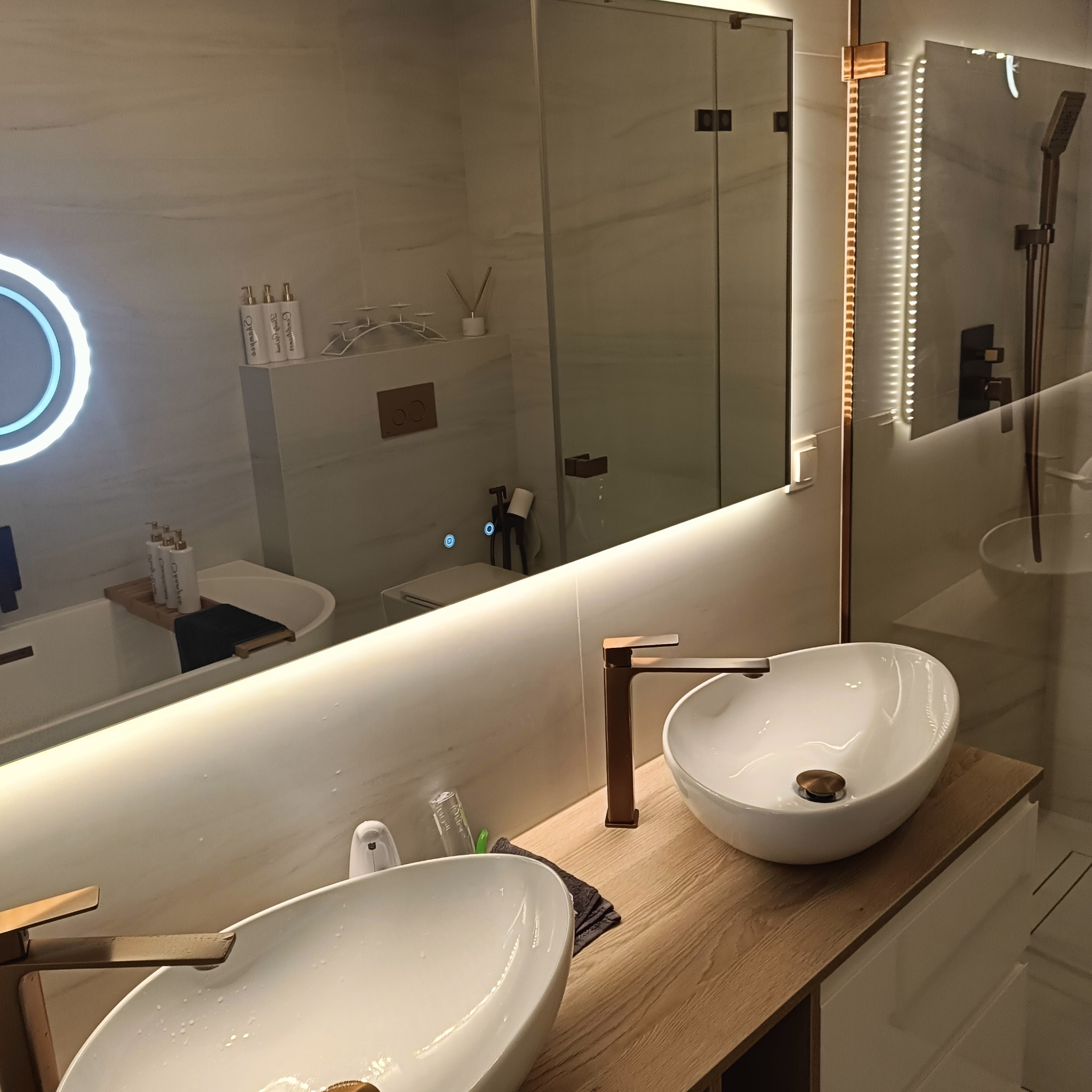 Nowoczesna łazienka z dwoma umywalkami wpuszczanymi w drewniany blat, lustro z podświetleniem LED i złotą armaturą. Widoczna wanna i kabina prysznicowa.