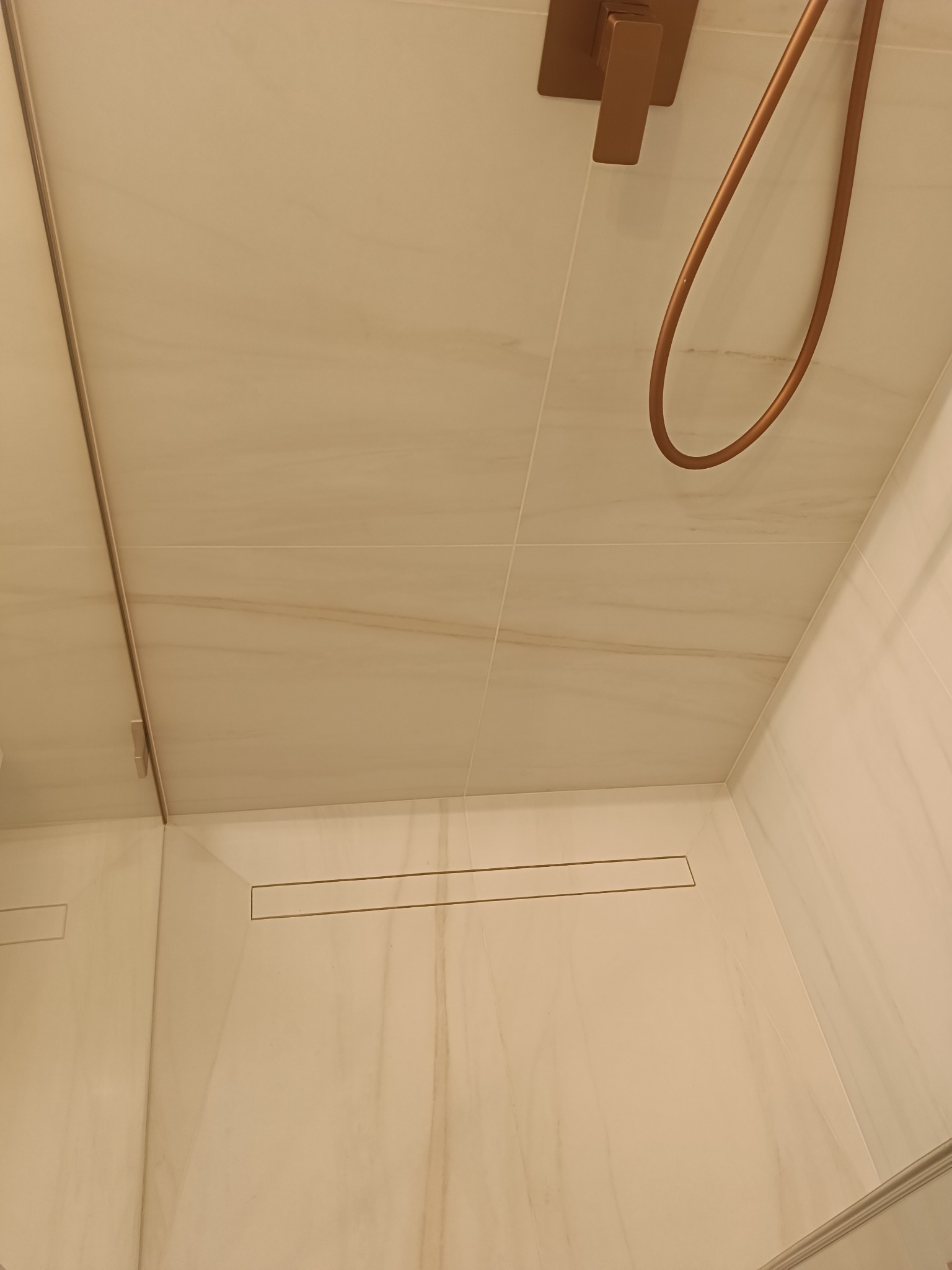 Beżowa kabina prysznicowa z odpływem liniowym i miedzianą armaturą. Płytki imitujące marmur na ścianach i suficie. Minimalistyczny design.