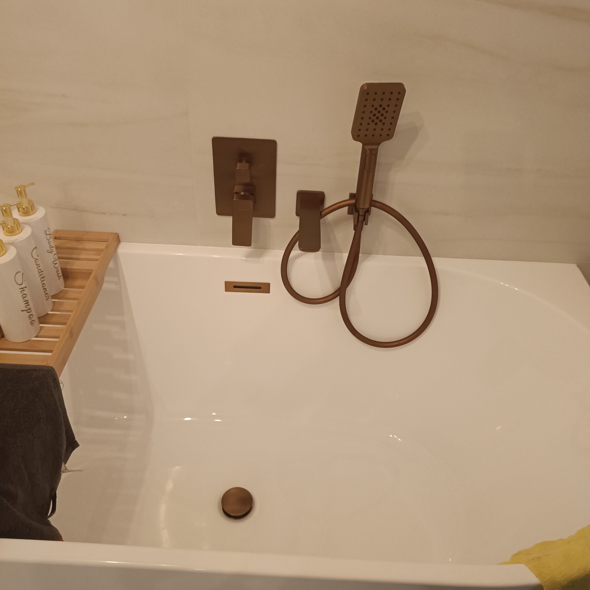 Biała wanna z brązową armaturą, w tym bateria, słuchawka prysznicowa i odpływ. Na wannie bambusowa półka z butelkami kosmetyków. Marmurowe płytki w tle.