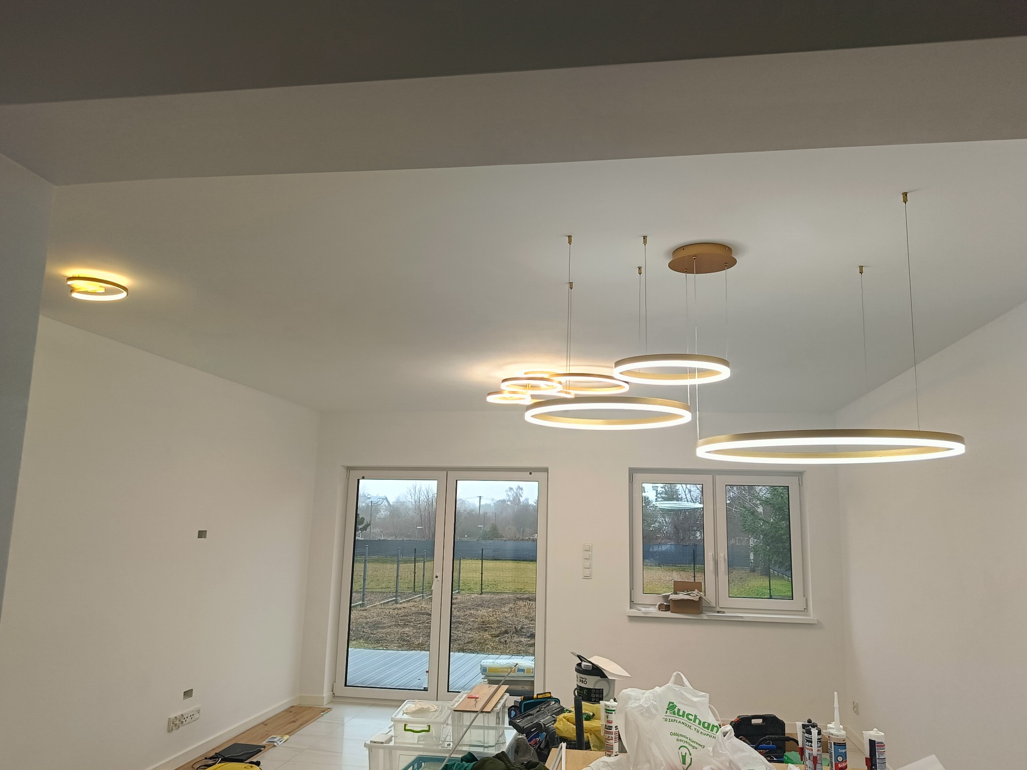 Nowoczesne oświetlenie LED w jasnym wnętrzu. Okrągłe lampy wiszące z ciepłym światłem, białe ściany i okna z widokiem na zewnątrz. Etap wykończenia, widoczne materiały budowlane.