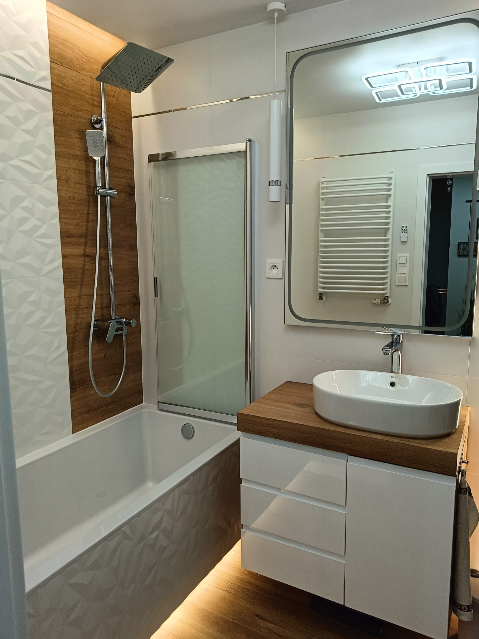 Nowoczesna łazienka z drewnianymi akcentami: kabina prysznicowa, umywalka na drewnianym blacie, lustro z podświetleniem i dekoracyjne płytki 3D.