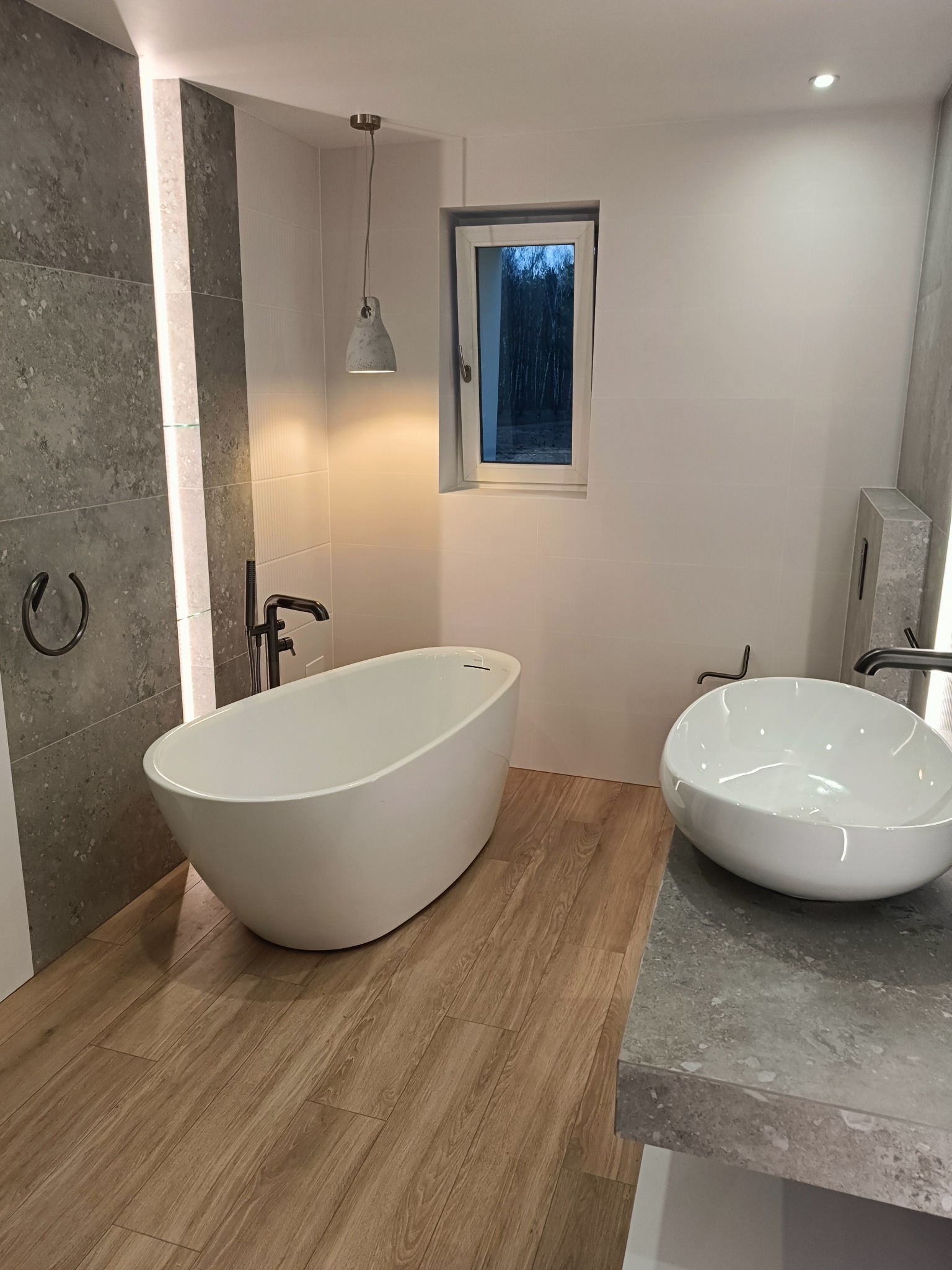 Nowoczesna łazienka z wolnostojącą wanną, umywalką nablatową i drewnopodobną podłogą. Szare płytki z oświetleniem LED. Okno z widokiem na zieleń.