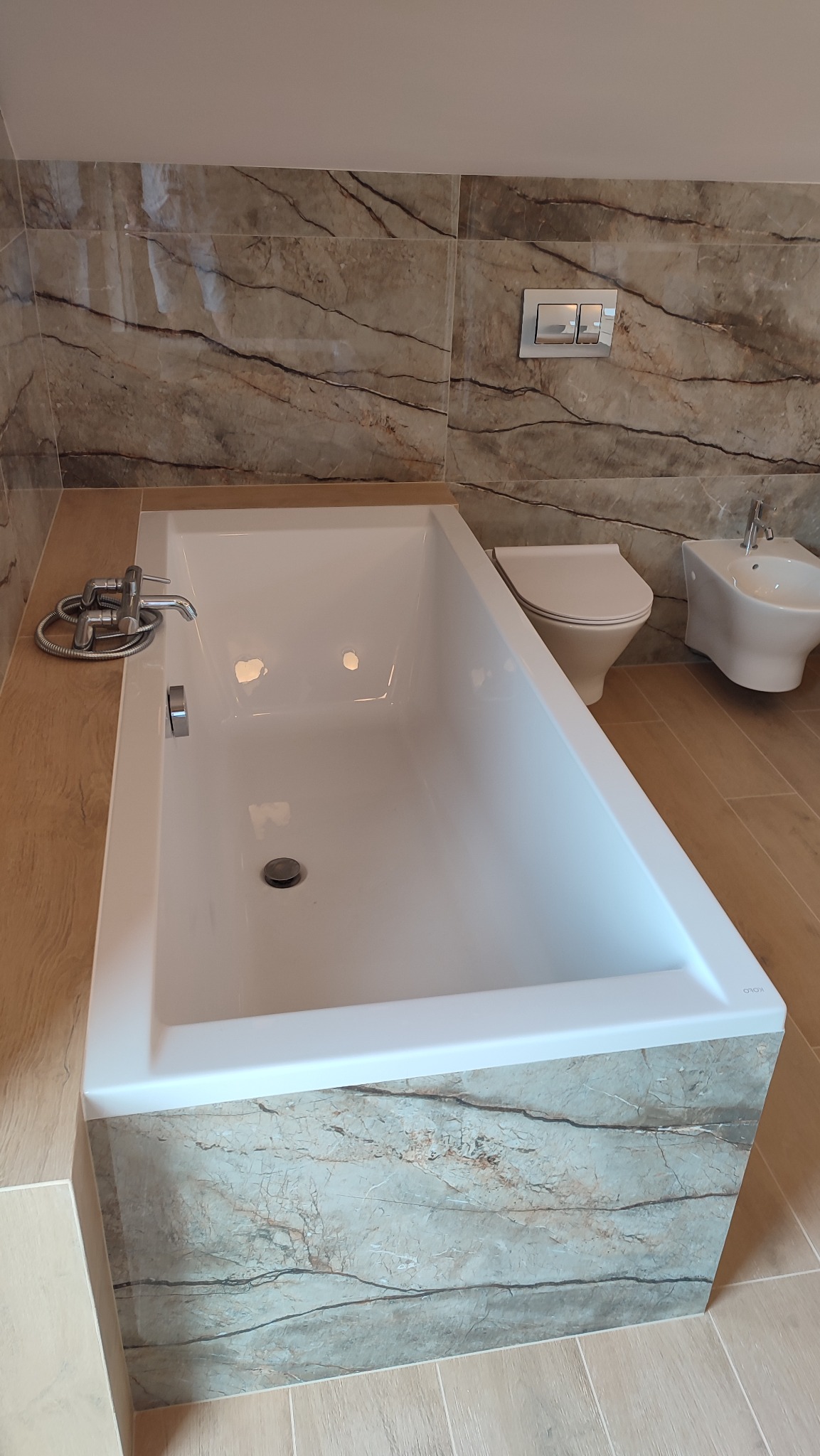 Wykończona łazienka z wanną obudowaną płytkami imitującymi marmur, toaleta i bidet w tle, całość utrzymana w beżowo-brązowej kolorystyce.