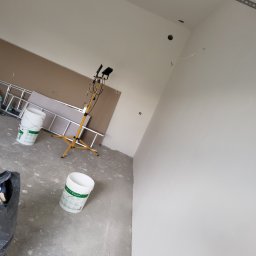 Adrian Sroka - Świeżo pomalowana ściana w jasnym pomieszczeniu z widocznymi śladami prac wykończeniowych, stojące wiadra z farbą, drabina i lampa budowlana.