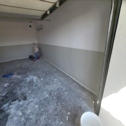 Adrian Sroka - Świeżo pomalowane wnętrze garażu z dwukolorową ścianą (szary dół, biała góra), widoczne puszki z farbą, drabina i plamy farby na betonowej podłodze, białe wiadro na pierwszym planie.