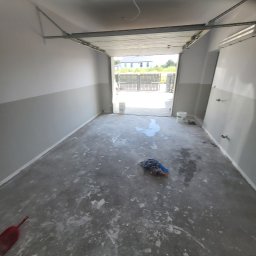 Adrian Sroka - Wnętrze garażu w trakcie malowania, dolna część ścian szara, podłoga z zabrudzeniami po farbie, widoczna szufelka i szmata.