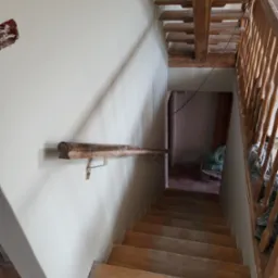 Klatka schodowa w trakcie remontu: pomalowane ściany na jasny kolor, drewniane schody i poręcz, widoczne ślady prac remontowych na podłodze, niezabezpieczone kable elektryczne.