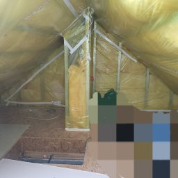 Adrian Sroka - Poddasze w trakcie ocieplania: żółta wełna mineralna zabezpieczona folią, drewniany szkielet konstrukcyjny, fragment podłogi z płyty OSB, widoczne przewody elektryczne i prowizoryczne oświetlenie.