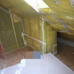 Poddasze w trakcie prac izolacyjnych, widoczna wełna mineralna zabezpieczona folią i taśmą, surowa podłoga z płyt OSB, fragment ściany z przytwierdzoną łatą.