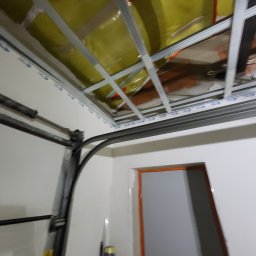 Adrian Sroka - Montaż metalowej konstrukcji pod sufit podwieszany z widoczną izolacją z wełny mineralnej, fragment ościeżnicy drzwiowej i prowadnicy bramy garażowej.