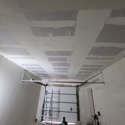 Adrian Sroka - Sufit garażu w trakcie przygotowania do malowania, widoczne płyty kartonowo-gipsowe i miejsca wypełnione masą szpachlową, drabina i brama garażowa w tle.