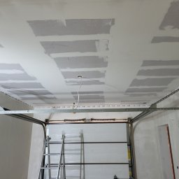 Adrian Sroka - Sufit w trakcie prac wykończeniowych z widocznymi fragmentami płyt kartonowo-gipsowych i nałożonymi pasami szpachli, drabina oparta o zamkniętą bramę garażową, widoczne przewody elektryczne...