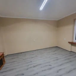 Pusty pokój z beżowymi ścianami, szarą podłogą imitującą drewno i drewnianą szafką RTV w rogu, oświetlony lampą sufitową i światłem z okna.